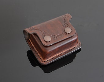 May include: A small, dark brown leather pouch with a flap secured by two metal snaps. The flap and top edge feature decorative embossed floral patterns. The pouch is rectangular with a stitched border, suggesting a handcrafted design.