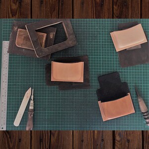 May include: A green cutting mat with four leather pieces, two tools, and a ruler. The leather pieces are different sizes and shapes, and some have a brown color. The tools are a wooden tool and a metal tool.