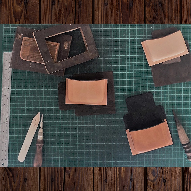 May include: A green cutting mat with four leather pieces, two tools, and a ruler. The leather pieces are different sizes and shapes, and some have a brown color. The tools are a wooden tool and a metal tool.