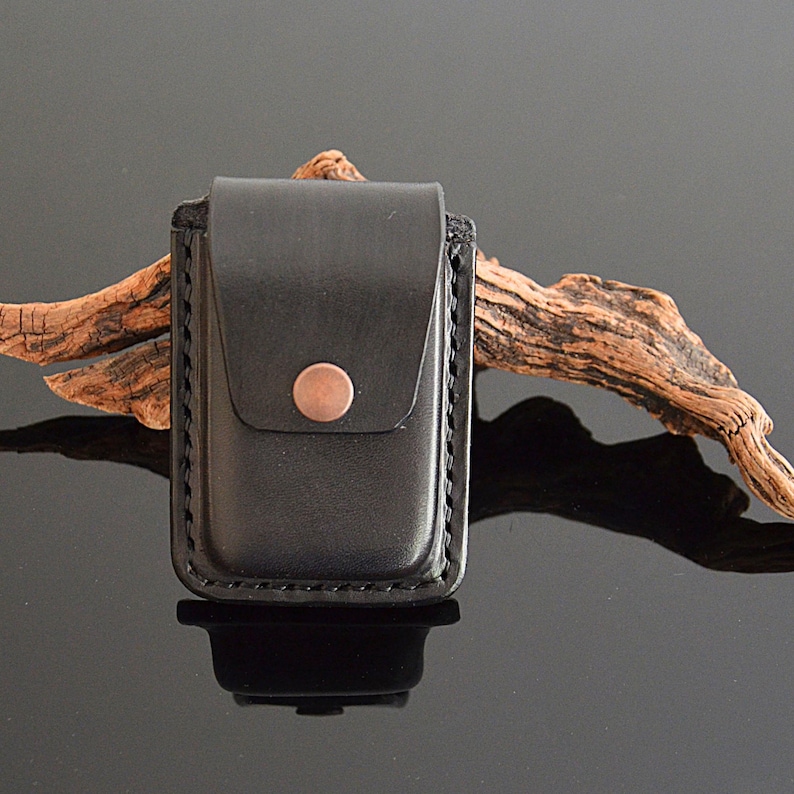 Puede incluir: Una funda de cuero negro con cierre de bot&oacute;n de cobre. La funda est&aacute; dise&ntilde;ada para guardar un objeto peque&ntilde;o, como un encendedor o una navaja de bolsillo.