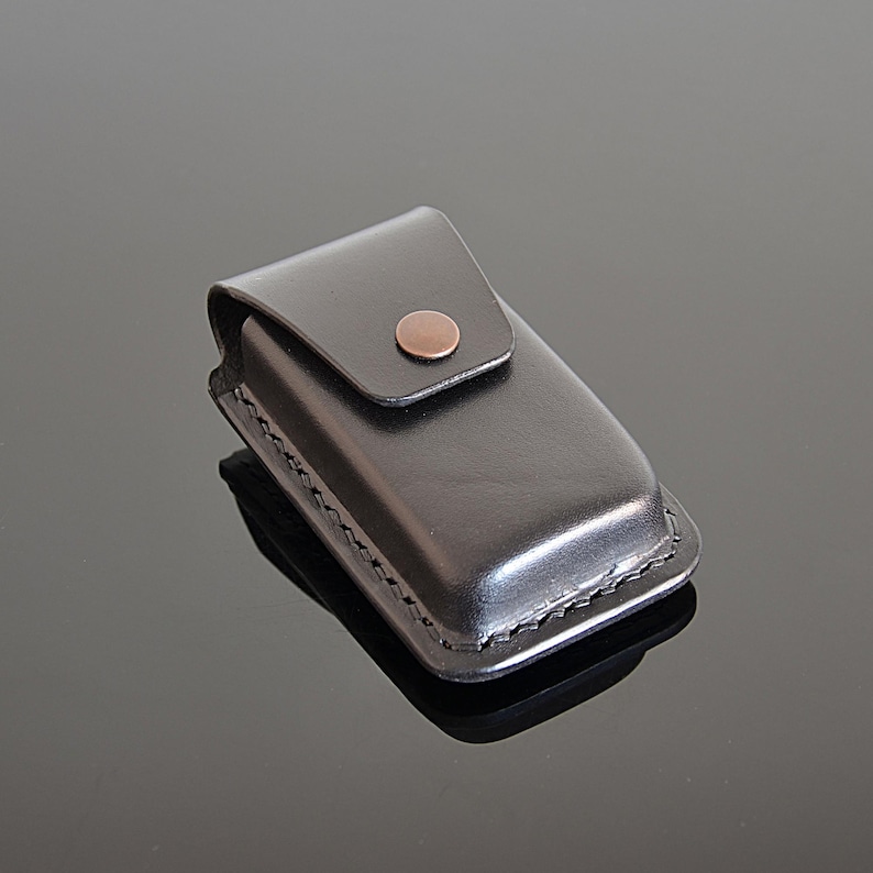 Puede incluir: Una funda de cuero negro con una solapa y un cierre de bot&oacute;n de cobre. La funda est&aacute; dise&ntilde;ada para contener un objeto peque&ntilde;o, como un encendedor o una navaja de bolsillo.