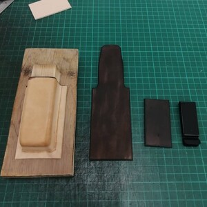 Peut inclure: Ensemble d'outils et de composants de maroquinerie. Comprend un moule en bois, une pi&egrave;ce de cuir marron fonc&eacute;, un petit rectangle et un clip noir. Les articles sont dispos&eacute;s sur un tapis de coupe vert.