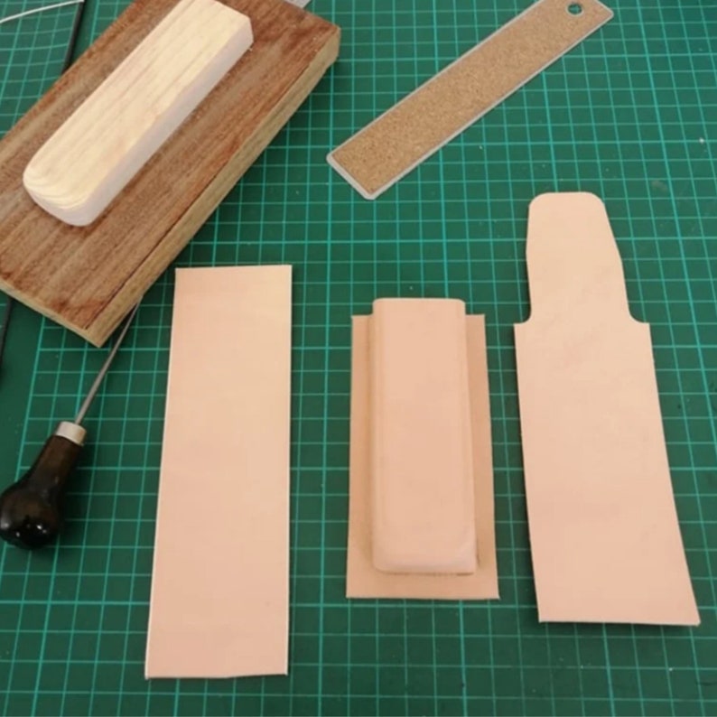 Peut inclure: Un espace de travail de maroquinerie avec divers outils et mat&eacute;riaux. Comprend un maillet en bois, une r&egrave;gle &agrave; dos de li&egrave;ge et plusieurs morceaux de cuir non finis de diff&eacute;rentes formes. Le fond est un tapis de coupe vert.