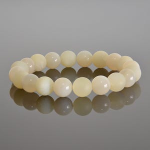 May include: A cream-colored beaded bracelet. The bracelet is made of round, smooth beads. The beads are strung together to form a circular shape. The bracelet is resting on a reflective surface.