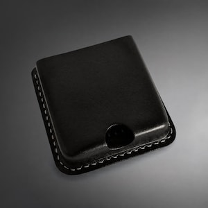 Könnte beinhalten: Ein schwarzer Kartenhalter aus Leder mit glatter, rechteckiger Form und abgerundeten Ecken. Die Kanten sind mit dicken, sichtbaren weißen Nähten versehen, die einen Kontrast zum dunklen Material bilden. Unten befindet sich ein kleiner Ausschnitt.
