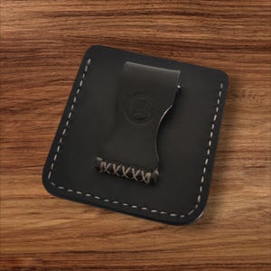 May include: A black leather money clip wallet with white stitching around the edges. The clip has a stamped logo and is attached to the wallet with a hand-stitched design. The wallet is square and sits on a wooden surface.