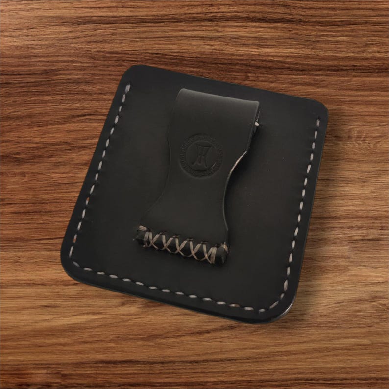 May include: A black leather money clip wallet with white stitching around the edges. The clip has a stamped logo and is attached to the wallet with a hand-stitched design. The wallet is square and sits on a wooden surface.