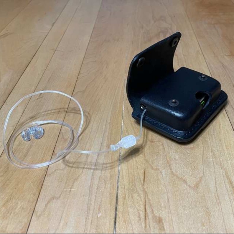May include: A black leather case with a clear tube and two small clear earpieces. The case has a flap that opens to reveal a small compartment.