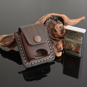 May include: A brown leather lighter case with a white stitch detail and a metal button. The case is sitting on a piece of driftwood next to a silver lighter with a skull and wings design and the text "Authentic Old School Rides".