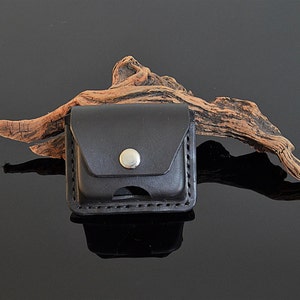 May include: A black leather case with a silver button closure. The case is sitting on a piece of driftwood.
