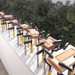 Peut inclure: Plusieurs coques de t&eacute;l&eacute;phone en bois, serr&eacute;es par des pinces noires et jaunes, avec un insert en cuir marron clair. Les coques sont en cours d'assemblage. Arri&egrave;re-plan de feuillage vert.