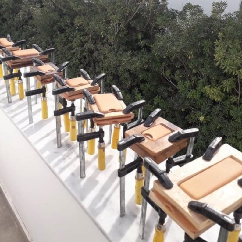 Peut inclure: Plusieurs coques de t&eacute;l&eacute;phone en bois, serr&eacute;es par des pinces noires et jaunes, avec un insert en cuir marron clair. Les coques sont en cours d'assemblage. Arri&egrave;re-plan de feuillage vert.