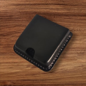 May include: A black leather wallet with a square shape and a curved cutout. The wallet has white stitching around the edges. The product is displayed on a wooden surface.