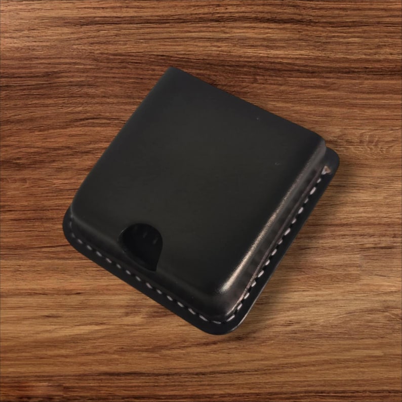 May include: A black leather wallet with a square shape and a curved cutout. The wallet has white stitching around the edges. The product is displayed on a wooden surface.