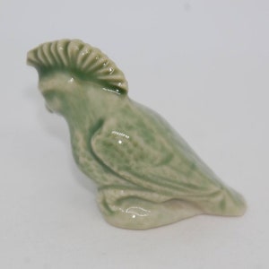 May include: A small ceramic figurine of a green bird with a spiky crest. The bird is sitting on its feet with its head turned to the side.