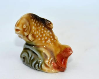 Może przedstawiać: Mała ceramiczna figurka brązowej i pomarańczowej ryby z zielono-białą podstawą.