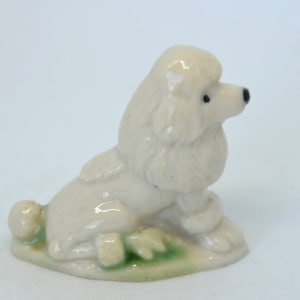 May include: A white porcelain figurine of a poodle sitting on a green base. The poodle has a fluffy tail and is looking to the right.