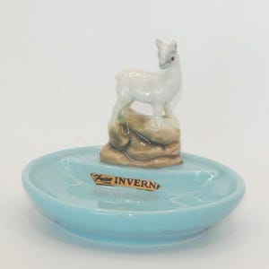 May include: A light blue ceramic trinket dish featuring a small white deer figurine standing on a brown rock formation. A rectangular sticker with the text "Frae INVERN" is attached to the centre of the dish, positioned in front of the deer.