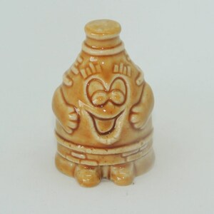 May include: A ceramic figurine of a smiling brown bottle with a cartoon face and arms. The bottle is wearing a brown belt and has a brown cap.
