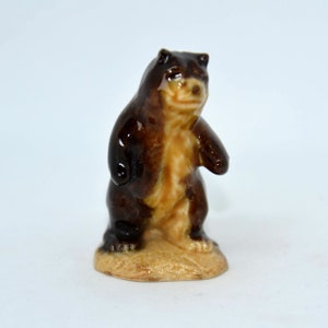 May include: A brown and tan ceramic figurine of a bear standing on a base. The bear has a brown body and a tan face and paws.