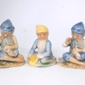 May include: Three ceramic figurines of gnomes, each wearing a blue hat and sitting on a base. The gnome on the left is holding a pipe, the gnome in the middle is holding a yellow object, and the gnome on the right is holding a small object.