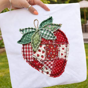 Puede incluir: Cojín blanco con un gran diseño de fresa aplicado. La fresa está hecha de tela roja con lunares blancos y parches de cuadros rojos y patrones florales. Hojas verdes y un tallo marrón completan el diseño.