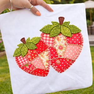 May include: White pillowcase featuring two appliqued strawberries. The strawberries are red with various patterned patches, green leaves, and brown stems. The pillowcase is held up by a person.