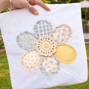 Puede incluir: Cojín blanco con un gran diseño de flor aplicada. La flor presenta pétalos en varios patrones: cuadros azules, estampados florales y lunares. Un tapete de ganchillo forma el centro de la flor, y también se incluye un pétalo amarillo.