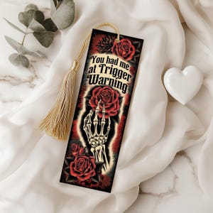 May include: A bookmark with a dark red and black design featuring roses and a skeleton hand holding a rose. The text "You had me at Trigger Warning" is displayed in white. A gold tassel hangs from the top.