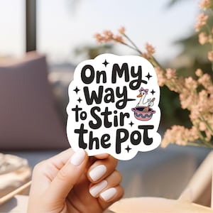 May include: A white sticker with the text "On My Way to Stir the Pot" in a bold, black font. The sticker features a cartoon goose wearing a pink bow and stirring a bowl. The sticker is held by a hand with white nail polish.