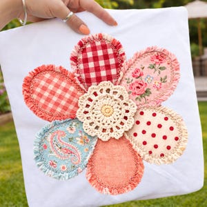 Puede incluir: Cojín blanco con un gran diseño floral. Los pétalos están hechos de varias telas estampadas, incluyendo cuadros rojos, estampados florales y lunares. Un tapete de ganchillo forma el centro de la flor.