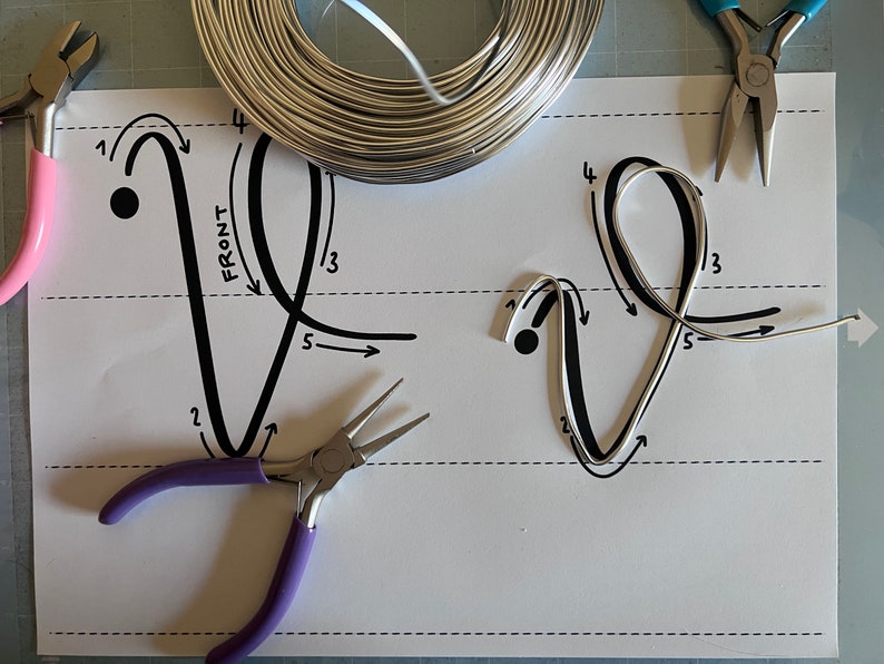 May include: A white sheet of paper with black lines and numbers, showing the steps for making a wire shape. Two pairs of pliers are on the paper, one pink and one purple. A roll of silver wire is in the upper left corner.
