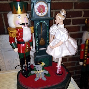 May include: A decorative Christmas display featuring a large nutcracker soldier in a red and green uniform, a ballerina doll in a white dress, and a green clock. A small gray mouse with a graduation cap is also present.