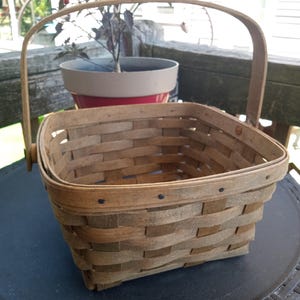 May include: A square, woven wooden basket with a handle. The basket is made of light brown wood and has a sturdy, rectangular shape. The handle arches over the top, and the basket is empty. A potted plant is in the background.
