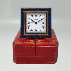 May include: A square Cartier desk clock with a white face, black Roman numerals, and black hands. The clock is set in a blue frame with gold accents and sits atop a red leather box with gold detailing.