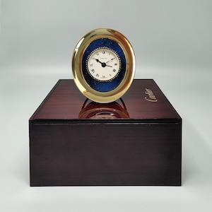 May include: A gold-toned oval clock with a white face and black Roman numerals. The clock has a blue textured background and sits on a dark wooden box. The word "Cartier" is visible on the box.