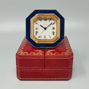 May include: A gold-toned octagonal clock with a white face and black Roman numerals, set within a blue frame. The clock is atop a red, leather-like box with gold accents. The clock face reads "Cartier".