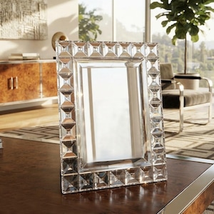 Pode incluir: Uma moldura retangular com uma borda de vidro transparente facetado com um padrão geométrico repetitivo em forma de pirâmide. A moldura possui um painel central de vidro liso e reflexivo e uma construção robusta para exibição em mesa.