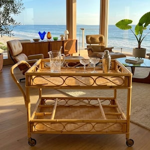 May include: A two-tiered bamboo bar cart with a handle and wheels. The top shelf holds a crystal pitcher, two coupe glasses, and a cocktail shaker. The cart is in a room with a view of the ocean.