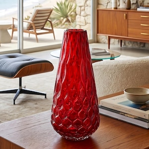 Può includere: Un vaso alto in vetro rosso vibrante con una silhouette affusolata e un motivo testurizzato a nido d'ape su tutta la superficie. Il vaso presenta una finitura lucida e una base ampia e arrotondata che si restringe verso l'apertura superiore.