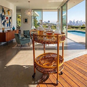 May include: A vintage rattan bar cart with two circular shelves. The top shelf holds several glass decanters filled with amber liquid. The cart is on wheels and has a woven design. The background shows a modern living room with a view of a city skyline.