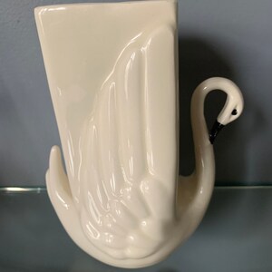 May include: White ceramic swan vase with a rectangular opening. The swan's neck is curved and the head is tilted to the side.