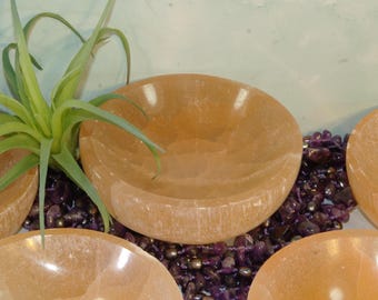 May include: Four small, round, orange-colored bowls made of selenite crystal. The bowls are arranged in a semi-circle on a bed of purple amethyst stones.