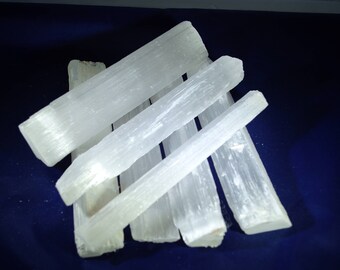 May include: A collection of translucent, white selenite crystal wands. The crystals are arranged in a pile, with varying lengths and textures. The background is a dark blue.
