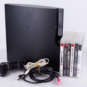 Puede incluir: Una consola PlayStation 3 negra con un mando negro y seis discos de juegos de vídeo. Los títulos de los juegos son "Fallout 3", "Call of Duty: Black Ops", "Lost Planet: Extreme Condition", "F1 2010", "Aliens vs Predator", y "Just Cause 2".