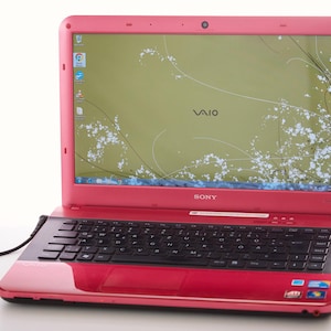 Puede incluir: Un portátil Sony VAIO rosa con un teclado negro y un touchpad negro. El portátil tiene un fondo de pantalla floral verde y blanco con la palabra "VAIO" en la pantalla.