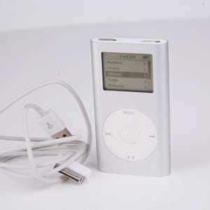 Puede incluir: Reproductor multimedia digital iPod plateado con cable de carga blanco. La pantalla muestra un menú con opciones como Listas de reproducción, Artistas y Canciones. La hora mostrada es 2:43 AM.