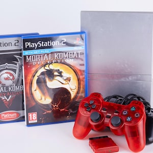 Könnte beinhalten: Eine PlayStation 2-Konsole mit zwei Spielhüllen, "Mortal Kombat: Deadly Alliance" und "Mortal Kombat: Deception." Ein roter Controller und eine Speicherkarte befinden sich vor der Konsole. Die Spielhüllen haben die Nummer 18.