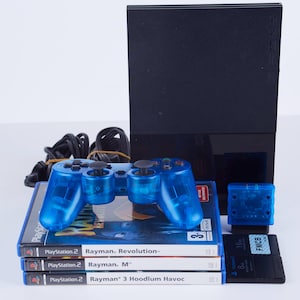 May include: A blue translucent PlayStation 2 controller with four PlayStation 2 game discs stacked on top of each other. The game discs are for Rayman Revolution, Rayman M, and Rayman 3 Hoodlum Havoc. A black PlayStation 2 console is in the background.