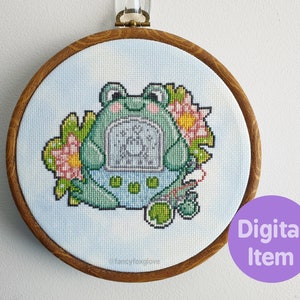 May include: Cross stitch pattern of a green frog holding a blue and white digital pet, surrounded by pink and yellow flowers. The pattern is framed in a wooden hoop.